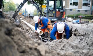 Twee medewerkers werken in een gegraven sleuf en plaatsen een leiding, met een graafmachine op de achtergrond