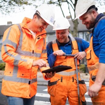 Drie medewerkers in oranje veiligheidskleding overleggen buiten bij de weg en kijken samen naar een tablet.