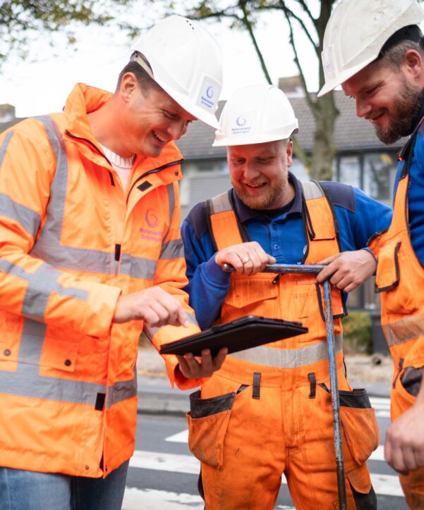 Drie medewerkers in oranje veiligheidskleding overleggen buiten bij de weg en kijken samen naar een tablet.